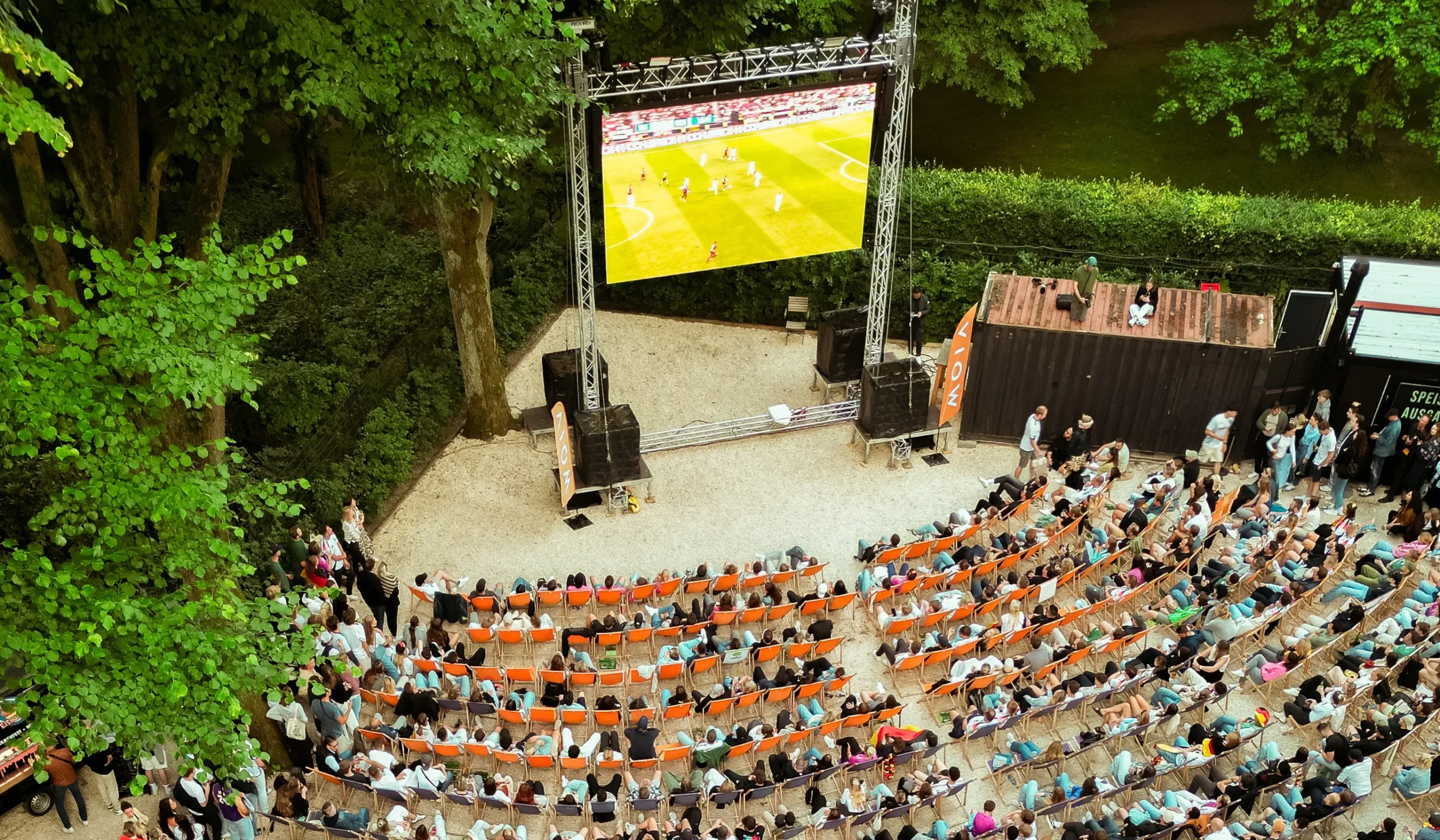 Große Zuschaueransammlung im Freien vor einer Leinwand, die ein Sportereignis zeigt, umgeben von Bäumen.