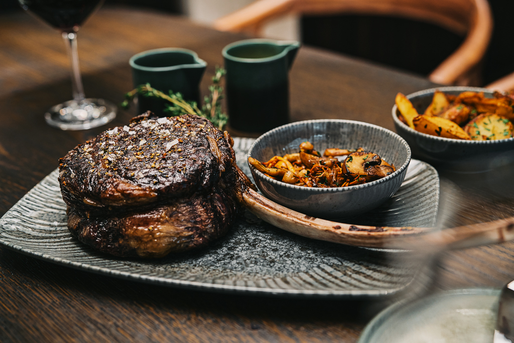 Saftiges Steak auf einem Teller, begleitet von sautierten Pilzen und gew&uuml;rzten Kartoffeln, mit Rotwein im Hintergrund.