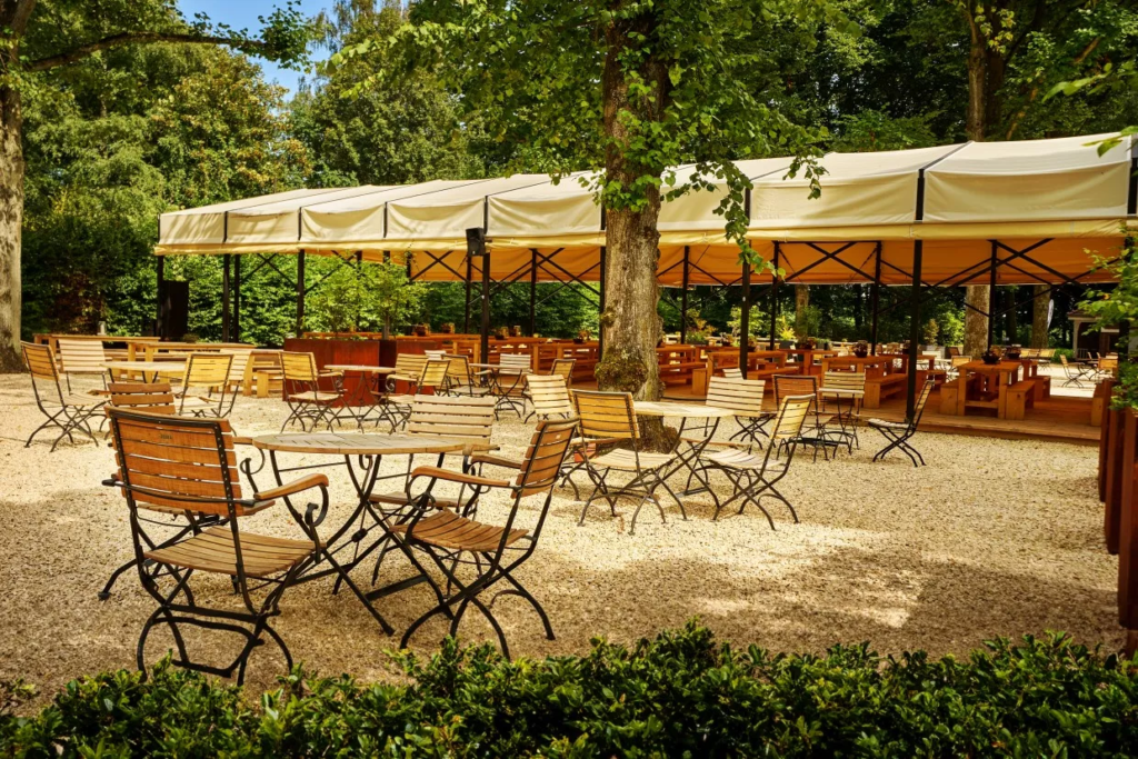 Blick auf ein Gartenrestaurant mit Holztischen und St&uuml;hlen unter einem wei&szlig;en Zelt, umgeben von B&auml;umen.