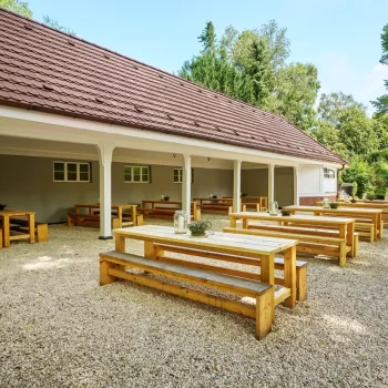 Holzbank-Tische im Freien unter einem &uuml;berdachten Bereich, umgeben von B&auml;umen und Kiesboden.