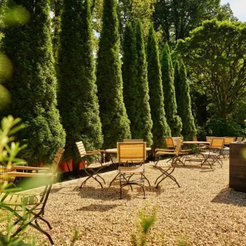 Ein Garten mit stehendem Kiesboden, Holztischen und St&uuml;hlen, umgeben von hohen, gr&uuml;nen Zypressen.