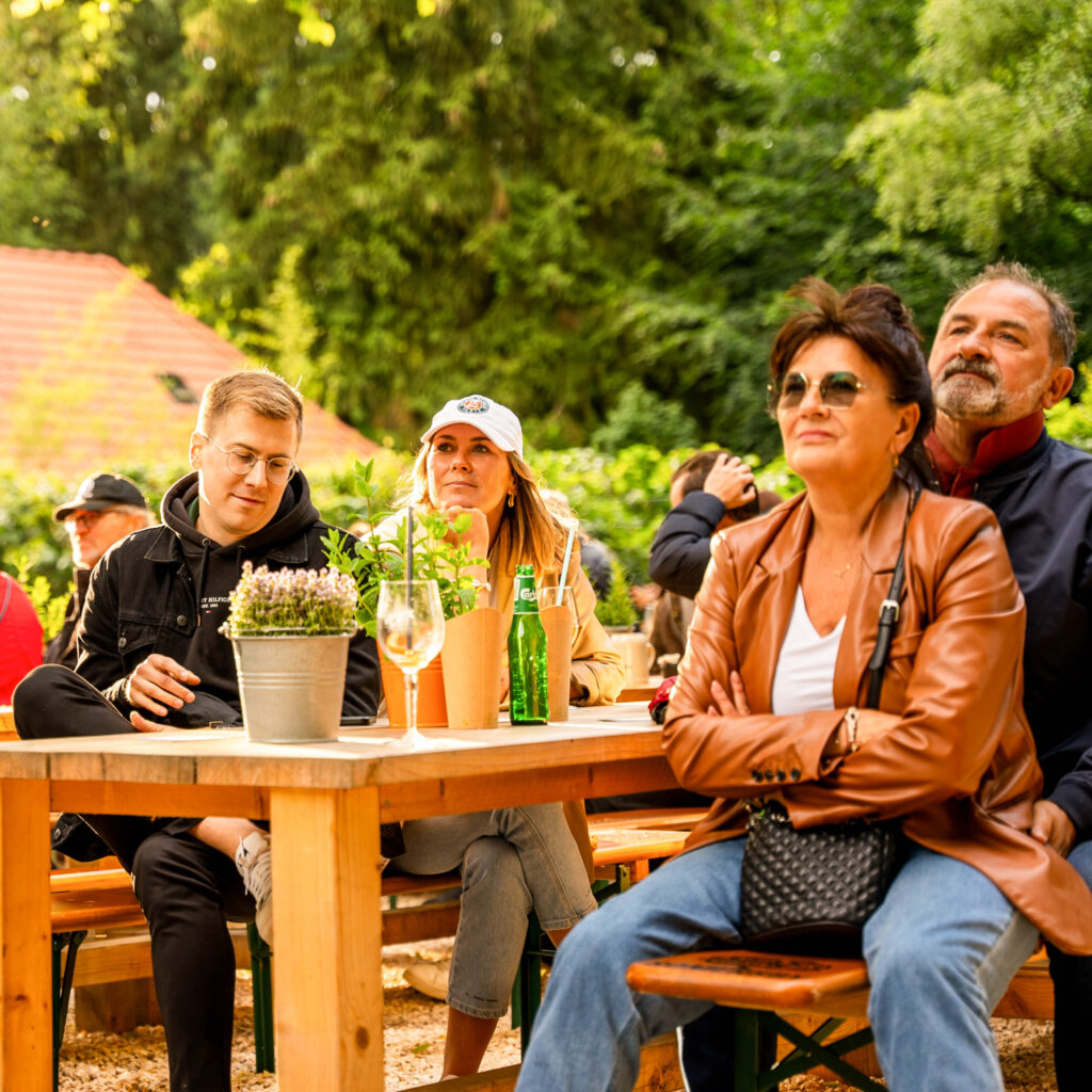 Eine Gruppe von vier Personen sitzt an einem Tisch im Freien, umgeben von Pflanzen und einer entspannten Atmosphäre.