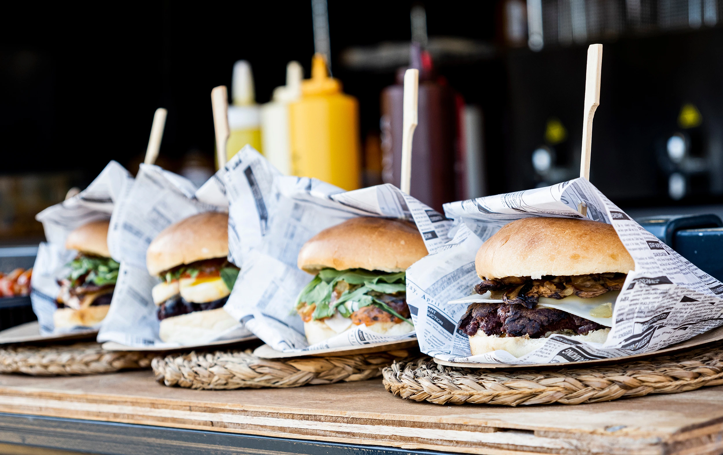 Vier Burger in Papier gewickelt, auf einem Holzbrett präsentiert, mit verschiedenen Belägen und Saucen im Hintergrund.