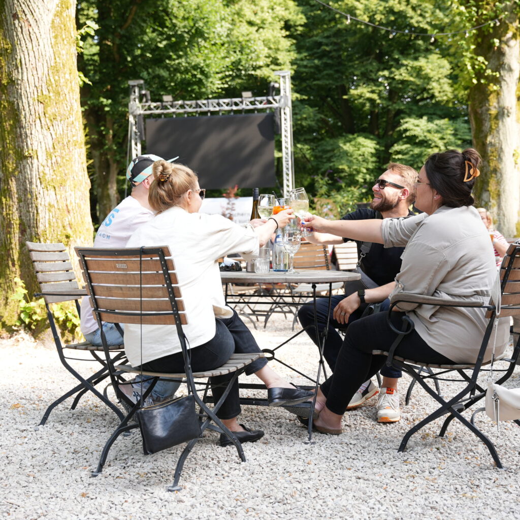 Gruppe von Menschen, die an einem Tisch im Freien anstoßen, umgeben von Bäumen und einer Bühne im Hintergrund.