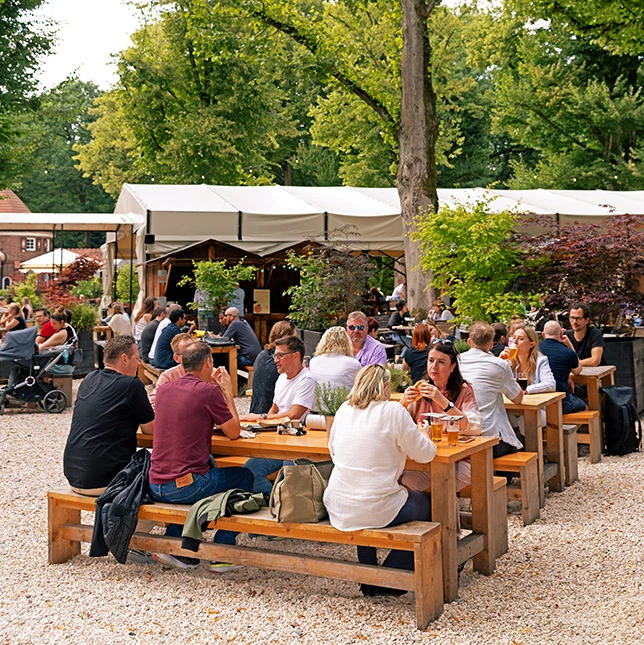 Menschen sitzen an Holztischen in einem Biergarten, umgeben von Bäumen und Pflanzen, mit einem Zelt im Hintergrund.