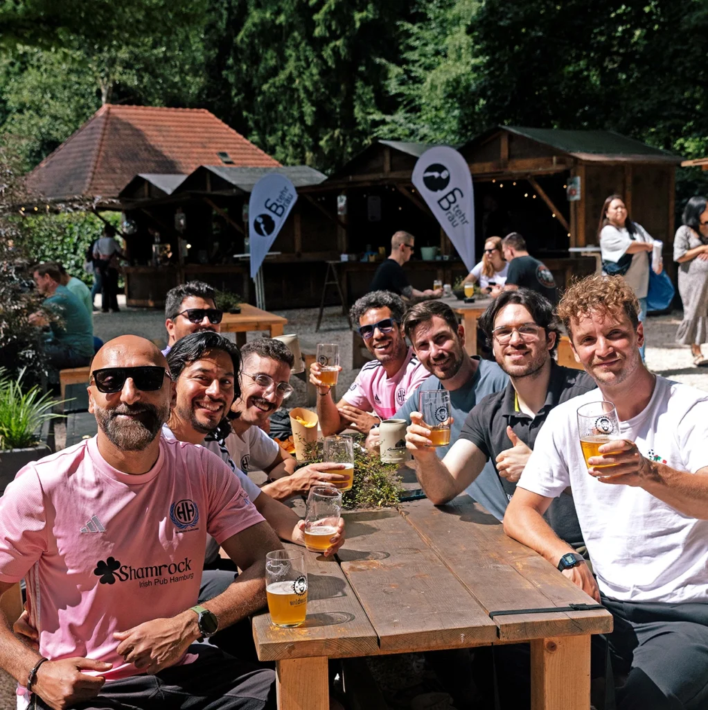 Gruppe von Männern in pinken Shirts, die an einem Tisch im Freien Bier trinken und lächeln, umgeben von Bäumen und Ständen.