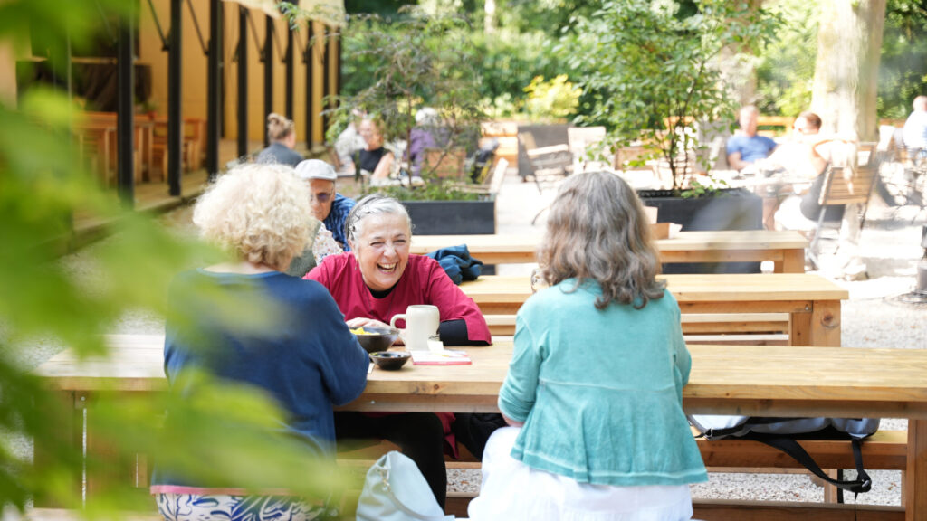 Drei Frauen sitzen an einem Tisch im Freien, eine von ihnen lacht, umgeben von Pflanzen und anderen Gästen im Hintergrund.