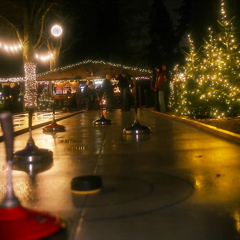 Eine beleuchtete Eisbahn mit Personen, umgeben von festlicher Dekoration und Tannenb&auml;umen im Dunkeln.