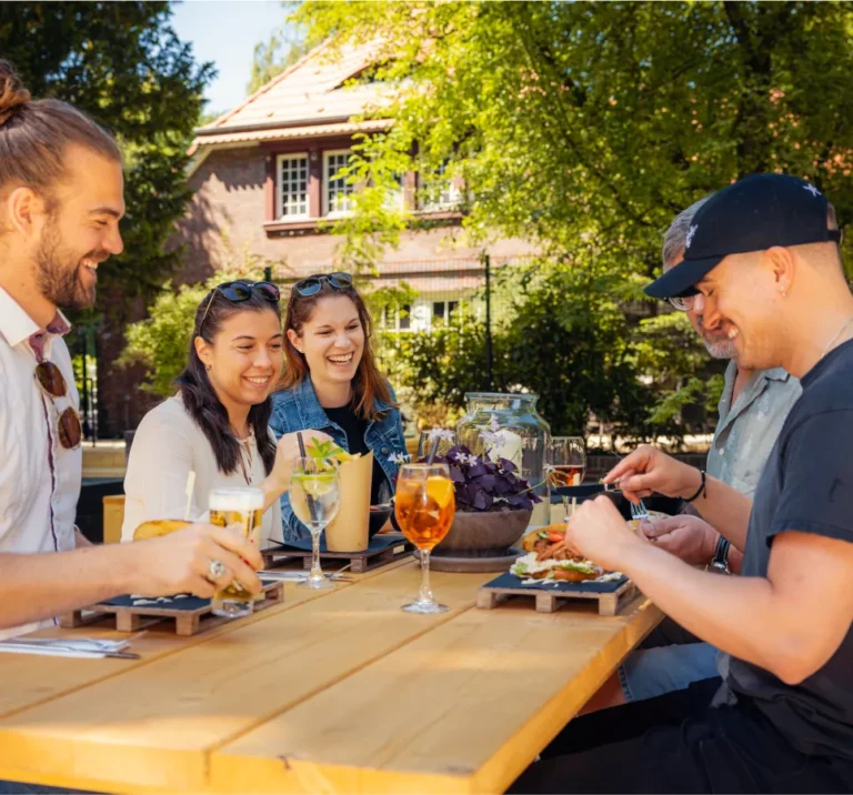 Fünf Personen sitzen an einem Tisch im Freien und genießen Essen und Getränke in einer sonnigen Umgebung.