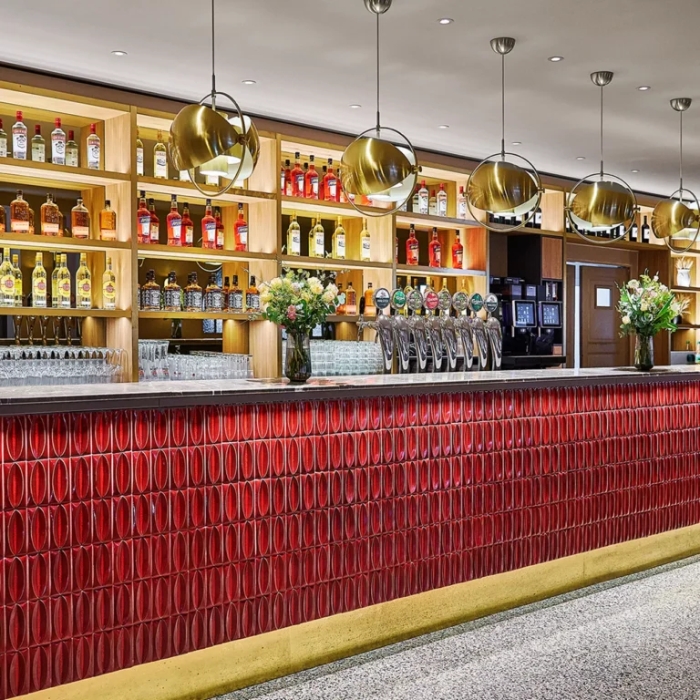 Moderne Bar mit Holzregalen, beleuchtetem Spirituosensortiment und eleganten, goldenen Pendelleuchten.