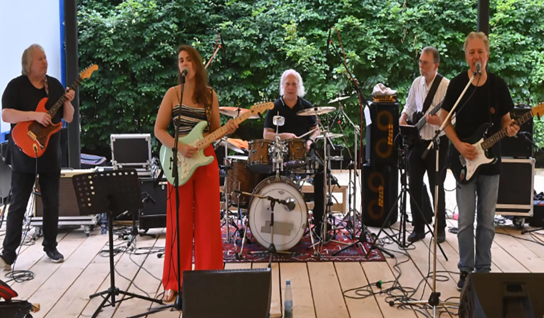 Eine Band spielt live auf einer Bühne, mit Sängerin, Gitarristen, Schlagzeuger und Keyboarder vor grünem Hintergrund.