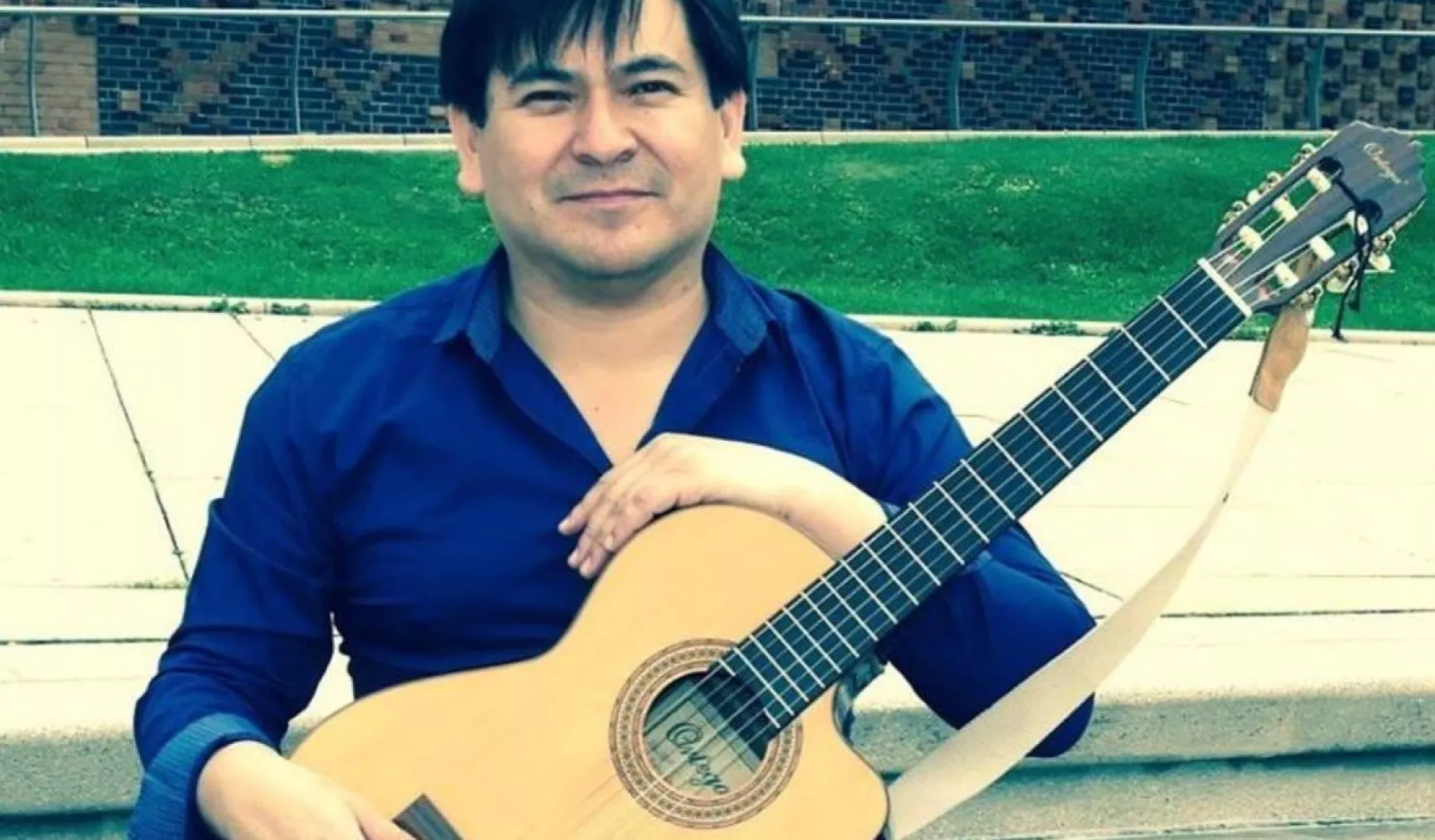 Mann mit Gitarre sitzt im Freien, trägt ein blaues Hemd und lächelt in die Kamera.