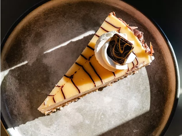 Ein St&uuml;ck K&auml;sekuchen auf einem grauen Teller, garniert mit Sahne und einer Schokoladendekoration.