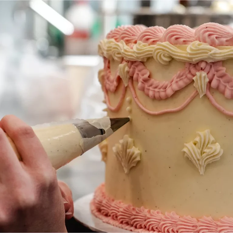 Eine Hand spritzt Zuckerguss auf eine dekorierte Torte mit rosa und cremefarbenem Muster.