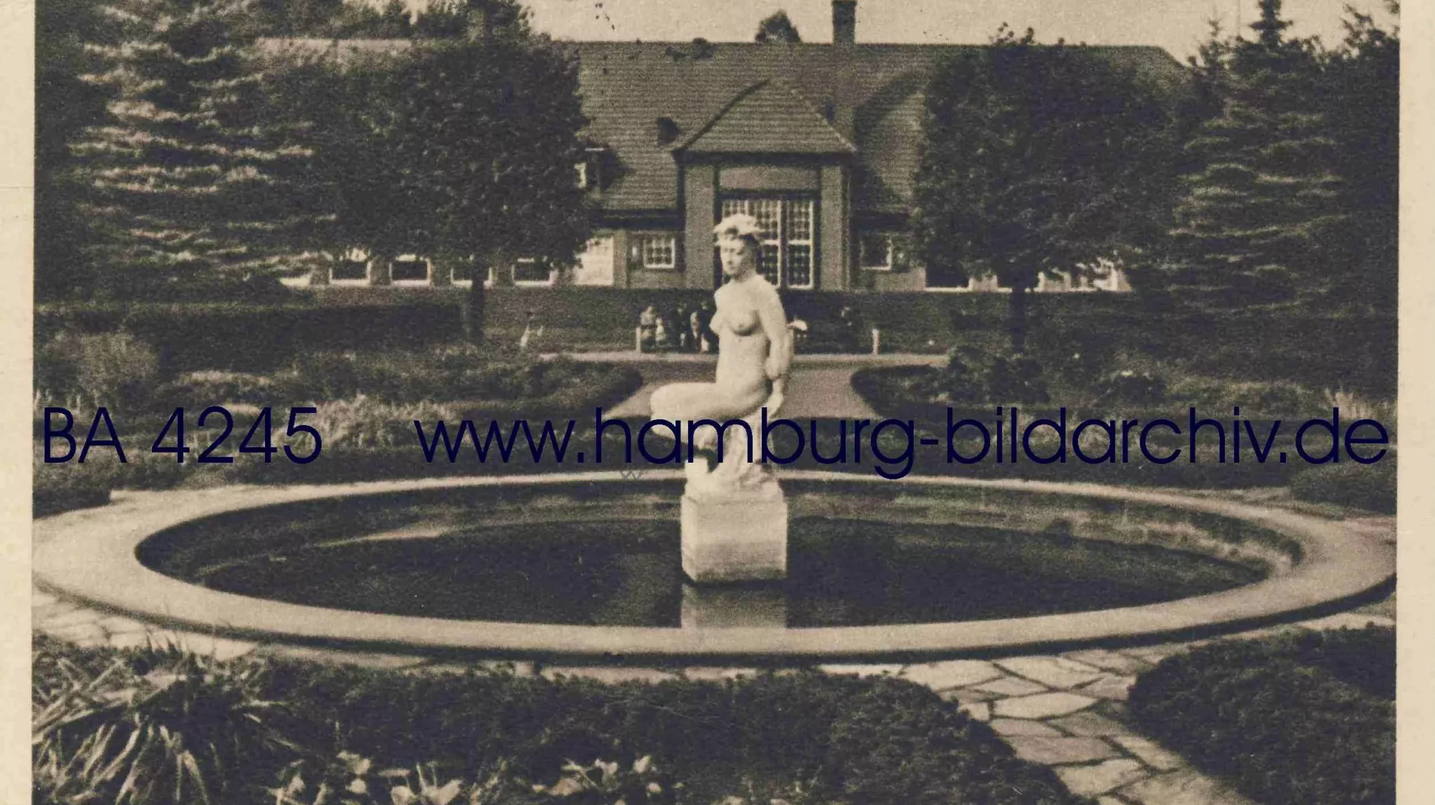 Eine Statue sitzt in einem Teich, umgeben von einem gepflegten Garten und einem historischen Geb&auml;ude im Hintergrund.