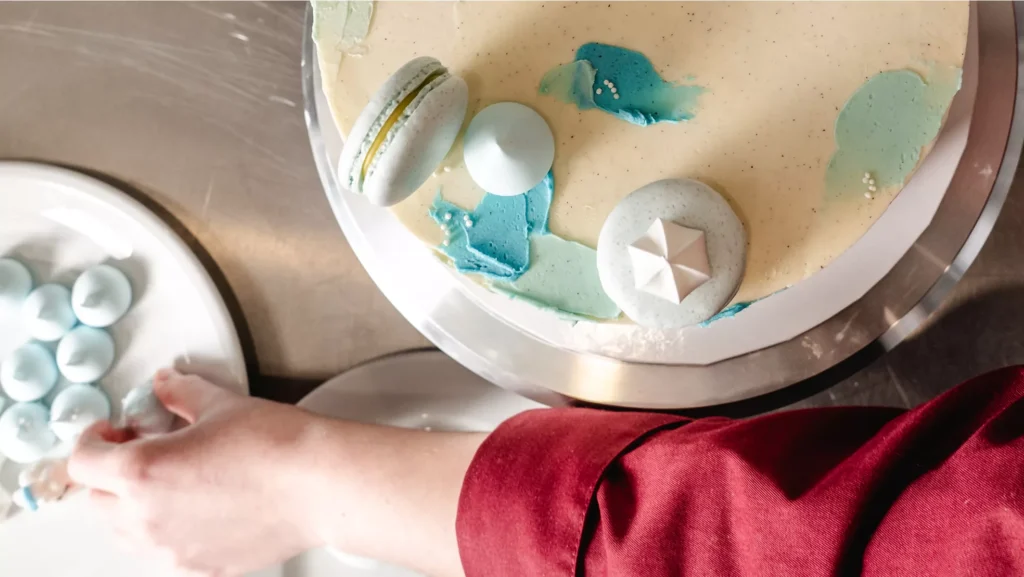 Eine Hand platziert kleine Macarons auf einem kunstvoll dekorierten Kuchen mit blauen Akzenten.