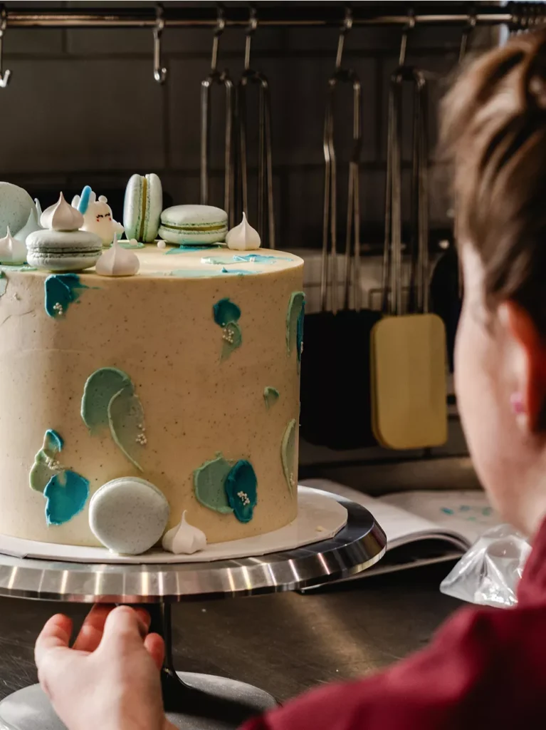 Ein Bäcker dekoriert eine mehrstöckige Torte mit blauen und grünen Akzenten sowie Macarons auf der Oberseite.