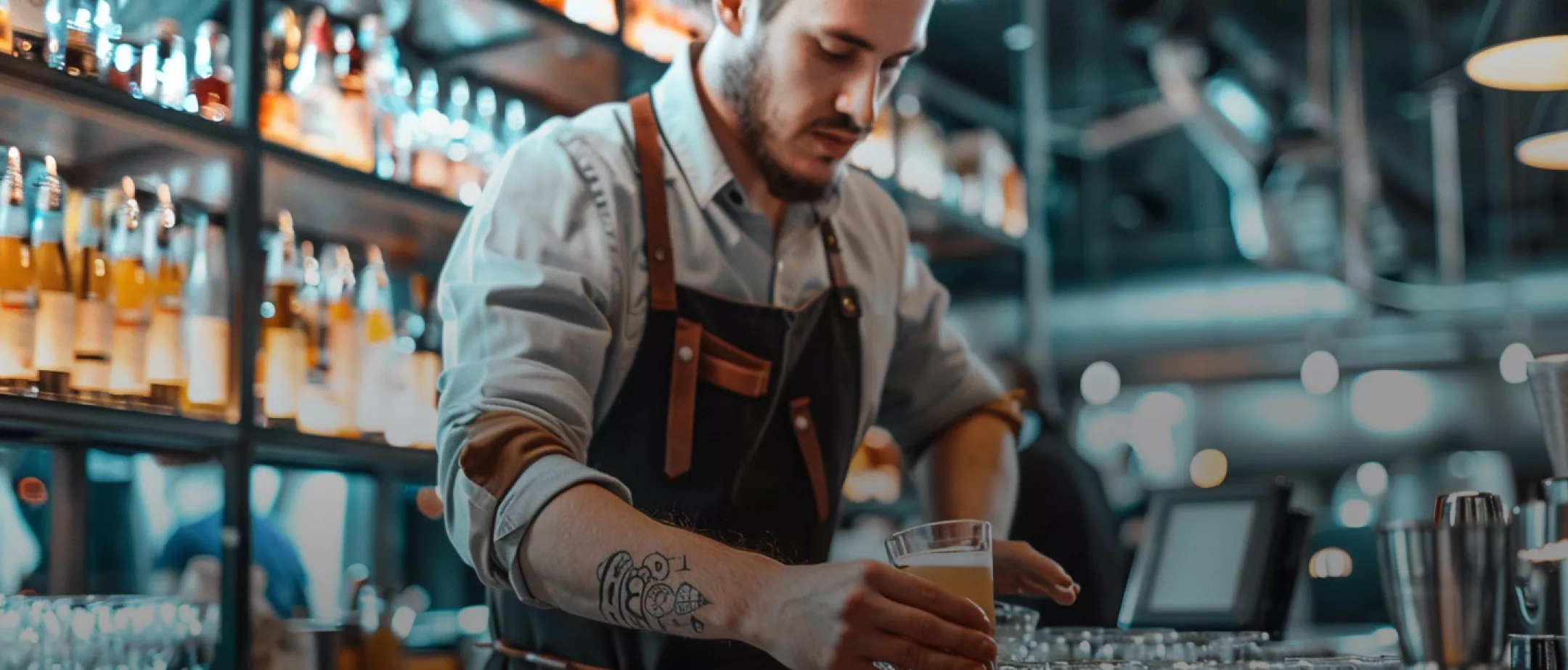 Ein Barkeeper mit t&auml;towiertem Arm serviert ein Getr&auml;nk an einer stilvollen Bar mit beleuchteten Flaschen im Hintergrund.