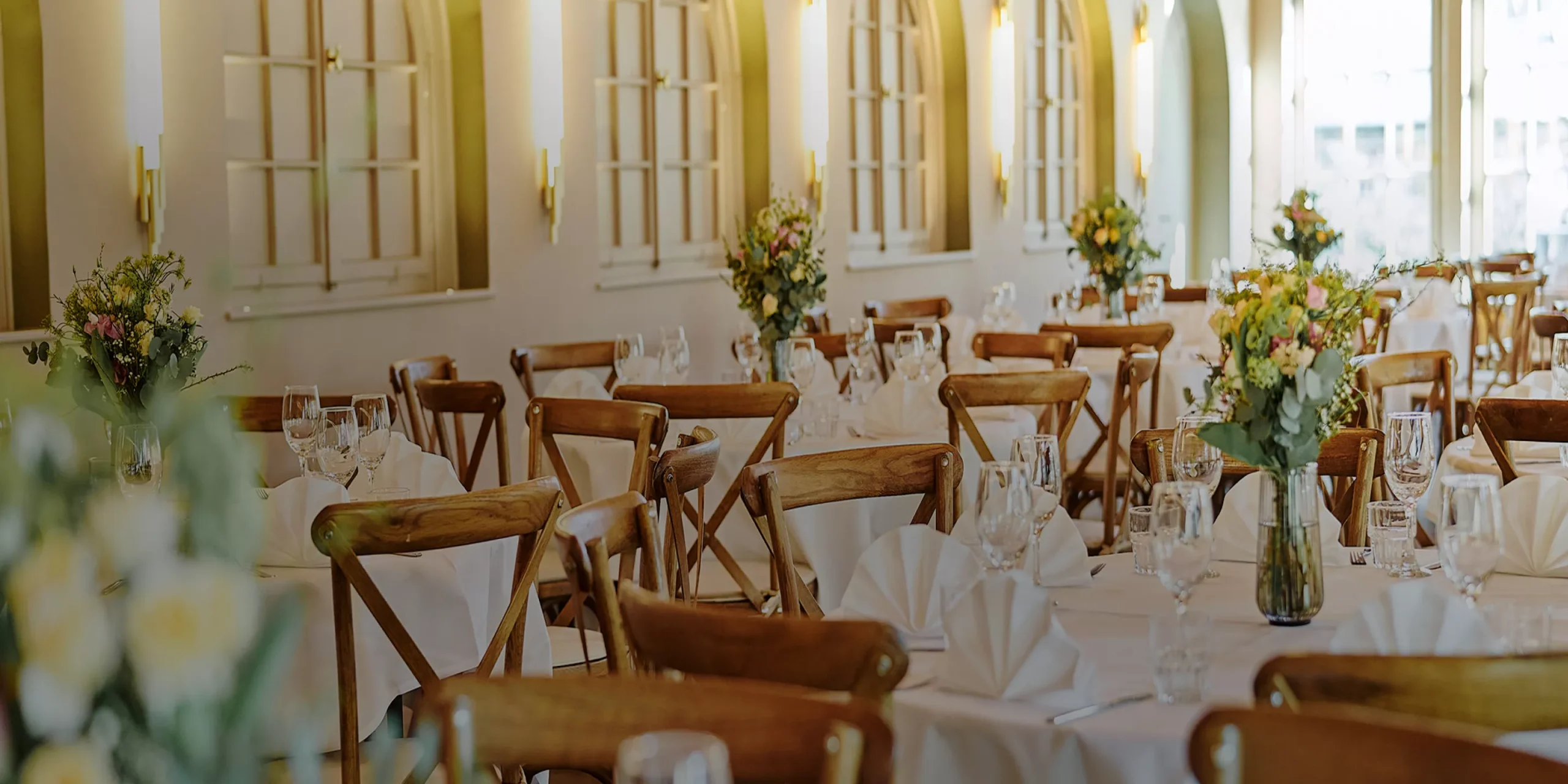 Eleganter Restaurantbereich mit Holztischen, wei&szlig;en Tischdecken und Blumendekorationen an den Fenstern.