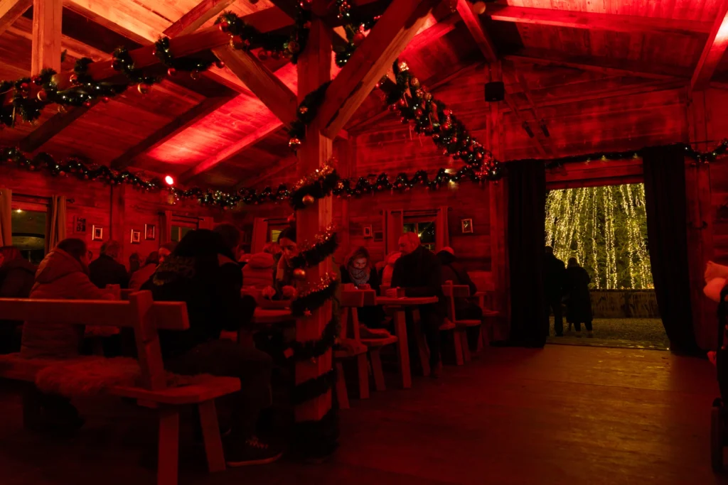 Gemütliche Holzhütte mit rotem Licht, festlich dekoriert, Menschen sitzen an Tischen, Blick auf beleuchtete Bäume draußen.