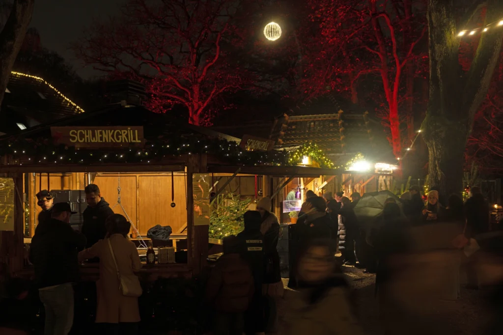 Ein festlicher Marktstand mit Menschen in der Nacht, umgeben von roten Lichtern und Weihnachtsdekoration.