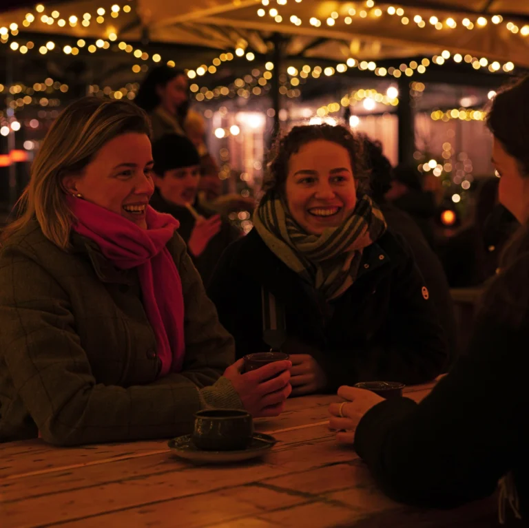 Drei Frauen sitzen an einem Tisch in einem festlich beleuchteten Au&szlig;enbereich und lachen miteinander.