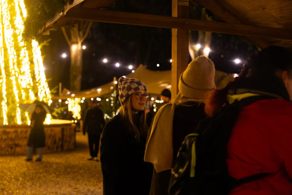 Eine Frau mit einer Mütze steht an einem festlich beleuchteten Stand auf einem Weihnachtsmarkt.