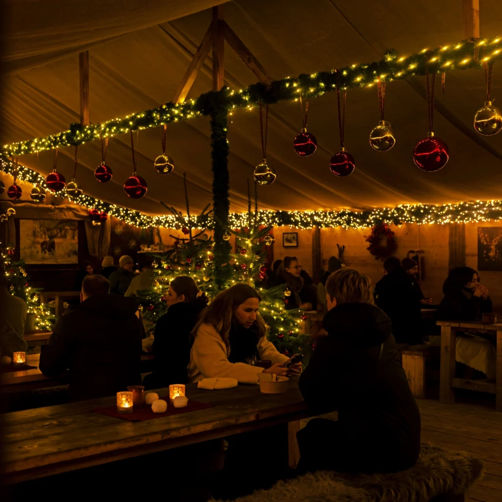 Eine weihnachtlich dekorierte Hütte mit Lichtern, Weihnachtsbaum und Gästen, die an Tischen sitzen und sich unterhalten.