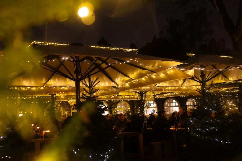 Beleuchteter Außenbereich eines Weihnachtsmarktes mit Gästen unter großen Sonnenschirmen und festlicher Dekoration.