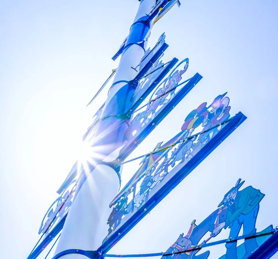 Ein farbenfroher Maibaum mit lebhaften Figuren, vor einem strahlend blauen Himmel, der in der Sonne gl&auml;nzt.