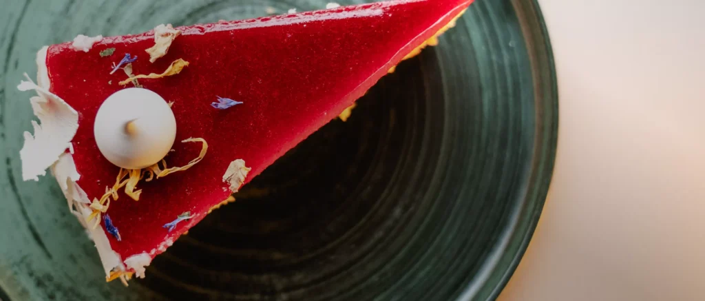 Ein Stück roter Kuchen auf einem dunklen Teller, dekoriert mit essbaren Blüten und einer kleinen weißen Kugel.