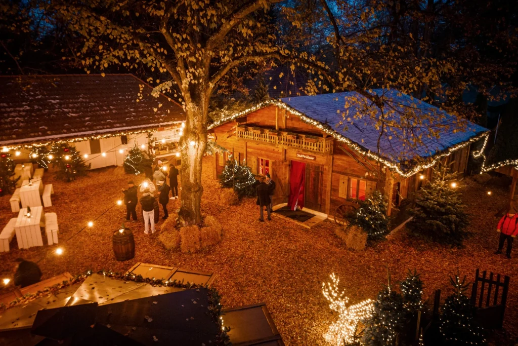Gemütliche Weihnachtsfeier im Freien mit festlich beleuchtetem Holzhaus und dekorierten Bäumen in herbstlicher Umgebung.