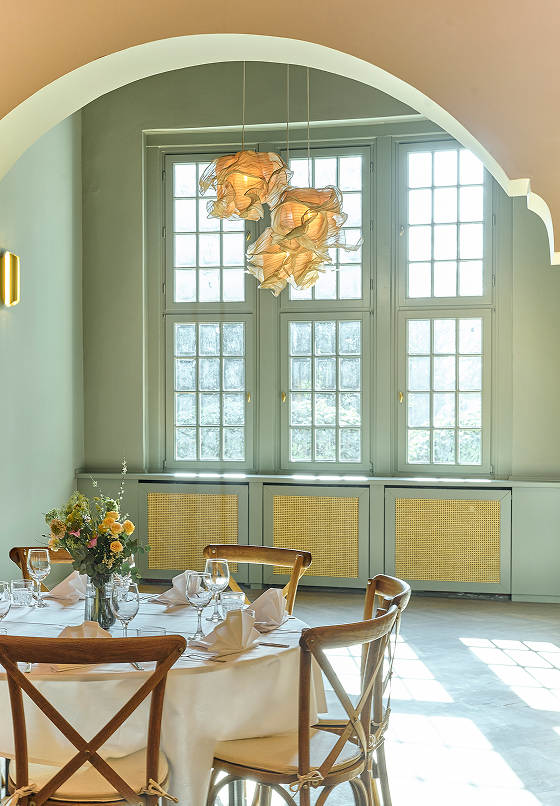 Eleganter Essbereich mit einem runden Tisch, dekoriert mit Blumen und stilvoller Beleuchtung unter einem hohen Fenster.
