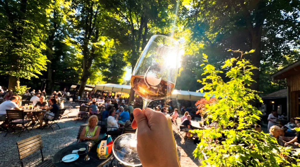 Ein Hand hält ein Weinglas mit Rosé, umgeben von einer lebhaften Gartenatmosphäre und Sonnenlicht durch Bäume.